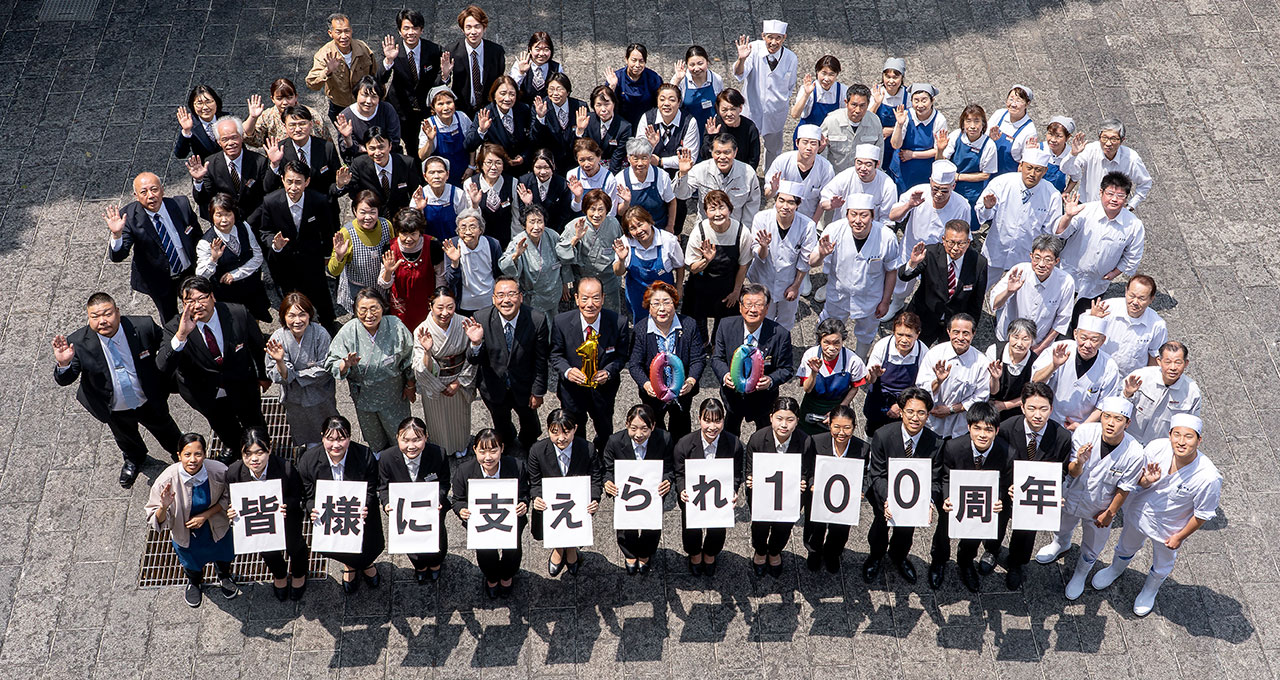 創業100周年を記念し、従業員が「皆様に支えられ100周年」と書かれたパネルを掲げて集合写真を撮っている様子。