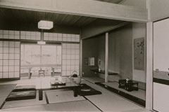 白黒写真。伝統的な日本家屋の室内を写している。畳が敷かれた部屋には、座卓と座布団が置かれ、落ち着いた空間。障子を開けると、縁側があり、その向こうには庭の風景が広がっている。